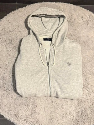 Ljusgrå zip hoodie Abercrombie & Fitch - Ljusgrå hoodie med dragkedja från Abercrombie & Fitch. Klassisk modell med huva och vita dragsnören! Riktigt snygg och trendig hoodie. Storlek L men passar lite som M. Skulle säga att den passar folk från 183-190cm! Hör av er vid frågor, pris går att diskuteras!🤩 