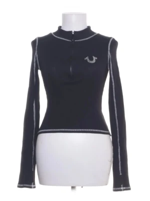 Svart långärmad topp från True Religion - Svart långärmad topp från True Religion med kontrasterande vita sömmar och halv dragkedja framtill. Toppen har en tight passform och en liten logga på bröstet som är silvrig. Perfekt för en trendig och sportig look. Har så att man kan ha tummen igenom ärmslutet.