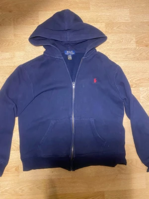 Marinblå hoodie från Polo Ralph Lauren - Snygg marinblå hoodie från Polo Ralph Lauren med dragkedja och huva. Klassisk röd logga broderad på bröstet och stora fickor framtill. Tillverkad i mjukt bomullsmaterial som är skönt att ha på sig. Perfekt för en avslappnad och stilren look.