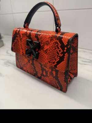 Röd handväska från Off-White - Säljer en riktigt snygg röd handväska från Off-White med svart ormmönster och det ikoniska Off-White-korset i svart på framsidan. Väskan har ett topphandtag och är gjord i ett glansigt material som liknar läder med reptilstruktur. Perfekt statement piece för dig som gillar att sticka ut. Använd mellan 5 till 8 tillfällen så den är som ny både på  in och utsidan. Kvitto har jag inte möjlighet att få fram så ni vet:)
