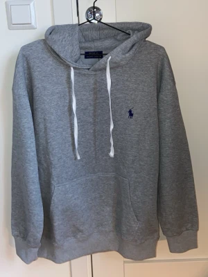 Polo Ralph Lauren grå hoodie HELT NY! - Snygg grå hoodie från Polo Ralph Lauren med klassisk blå logga broderad på bröstet. Hoodien har vit dragsko i huvan och en stor magficka framtill. Tillverkad i mjuk bomullsblandning och har ribbade muddar vid ärmar och nederkant. Hoodien är helt oanvänd!