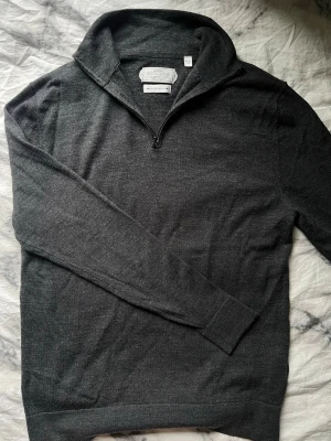 Mörkgrå merinoullströja med halvzip av These Glory Days - Snygg mörkgrå stickad tröja från These Glory Days i mjuk superior merinoull. Tröjan har halvzip, hög krage och långa ärmar. Perfekt för lager på lager och ger en clean look till både jeans och chinos.