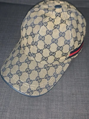 Gucci beige keps med GG-mönster - Säljer en beige keps från Gucci med klassiskt GG-monogram över hela kepsen. Kepsen har en böjd skärm och en snygg detalj med blå och röd rand på sidan. Tillverkad i canvasmaterial som ger en exklusiv känsla och streetig look.
