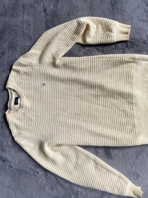 Beige  stickad tröja från Gant - Säljer en stilren beige stickad tröja från Gant med diskret logga på bröstet. Tröjan har ett strukturerat rutmönster, rund halsringning och långa ärmar. Perfekt för dig som gillar klassisk och enkel stil.