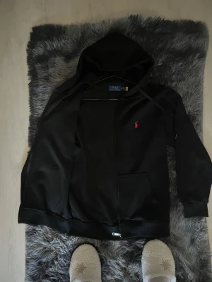 Svart hoodie från Polo Ralph Lauren - Säljer en svart hoodie från Polo Ralph Lauren med dragkedja framtill och två fickor. Klassisk röd logga broderad på bröstet. Huva med dragsko och ribbade muddar vid ärmar och nederkant. Perfekt för en chill och stilren look.