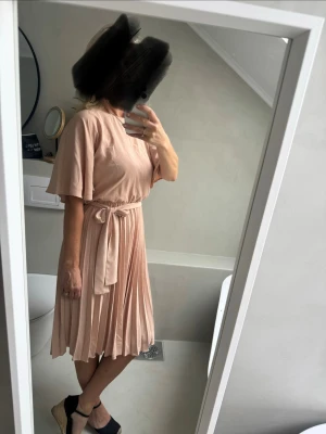 Ljusbeige omlottklänning med volang - Säljer en ljusbeige klänning med omlottknytning i midjan och vackra volangärmar. Kjolen är plisserad och klänningen har en mjuk, flowy känsla. Perfekt för dig som gillar en feminin och stilren look.