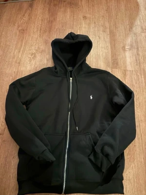 Svart Ralph Lauren Zip Hoodie  - Polo Ralph Lauren zip hoodie – svart – mycket fint skick Säljer en stilren svart hoodie från Polo Ralph Lauren med hel dragkedja och den klassiska vita broderade loggan på bröstet. Perfekt som vardagsplagg och enkel att matcha med det mesta. Färg: Svart Modell: Zip hoodie med huva Detaljer: Vit broderad logga på bröstet, kängurufickor framtill Skick: Mycket fint skick utan synliga hål eller större slitage Passform: Normal passform