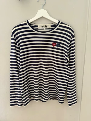 Comme des Garçons Randig M  - Randig långärmad tröja från Comme des Garçons PLAY med marinblå och vita ränder. På bröstet finns två broderade hjärtan, ett rött och ett blått, med ögon. Tröjan har rund hals och är i mjukt bomullsmaterial. Säljs pga för liten. Hör av er vid funderingar!