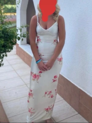Vit långklänning med blommor - Snygg vit långklänning med smala axelband och blommigt mönster i rosa och rött. Klänningen har en figurnära passform och v-ringad urringning, perfekt för sommarens alla tillfällen. Använd fåtal gånger men inga skador ser ut som ny.
