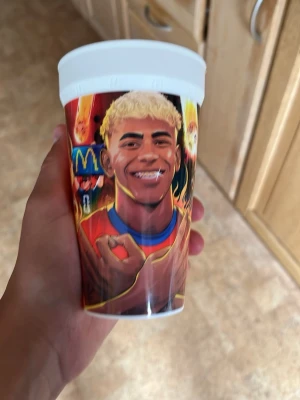 McDonald's plastmugg med fotbollstryck - Cool plastmugg från McDonald's med färgglad illustration av en fotbollsspelare i röd tröja och blont hår. Muggen har eldiga detaljer och sporttema runt om, perfekt för dig som gillar fotboll och samlarprylar.