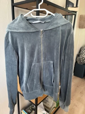 Grå velourhoodie från 157 - Mjuk och skön grå hoodie i velourmaterial från 157. Dragkedja framtill med rund metallring som detalj, två stora fickor och huva. Perfekt för en chill och avslappnad stil.