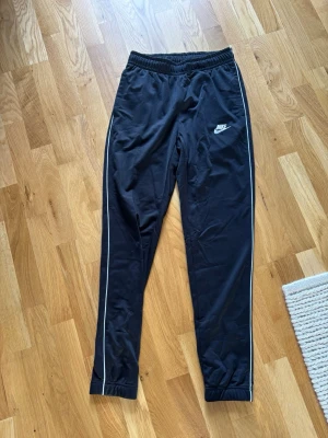 Svarta mjukis/vindbyxor från Nike - Snygga svarta mjukisbyxor från Nike med vita detaljer längs sidorna och klassisk logga på vänster ben. Storlek XS men kan även passa S om man är lite kortare. 