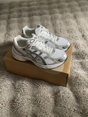 Asics 1130  - Snygga vita sneakers från Asics med silvriga detaljer och meshpaneler för extra ventilation. Klassisk låg modell med bekväm sula och tydlig logga på sidan. Perfekta för dig som gillar sportig stil och vill ha något som sticker ut lite extra.