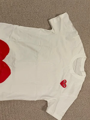 Vit t-shirt med rött hjärta - Vit t-shirt med ett ikoniskt rött hjärta med ögon tryckt på bröstet. Klassisk rund hals och korta ärmar. Perfekt för dig som gillar enkel men ändå lekfull design.