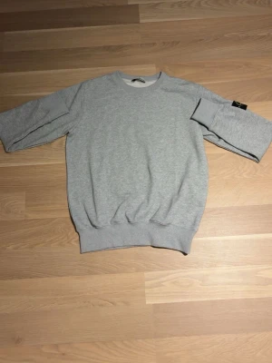 Grå sweatshirt från Stone Island - En stilren grå sweatshirt från Stone Island med klassisk rund hals och ribbade muddar. Tröjan har en ikonisk Stone Island-patch på vänster ärm och är gjord i mjukt bomullsmaterial. Perfekt för en avslappnad och trendig look.