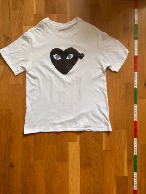 Vit Comme des Garçons t-shirt hjärta - Säljer en vit t-shirt från Comme des Garçons med det ikoniska svarta hjärtat med ögon tryckt på bröstet. T-shirten har rund halsringning och korta ärmar. Tillverkad i mjuk bomull för en skön känsla och clean look.