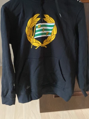 Svart Hammarby hoodie med logga - Svart hoodie från Hammarby IF med stor klubbemblem i gult, grönt och vitt på bröstet. Klassisk känguruficka framtill och huva med snörning. Perfekt för dig som vill visa ditt stöd för Bajen på ett stilrent sätt.