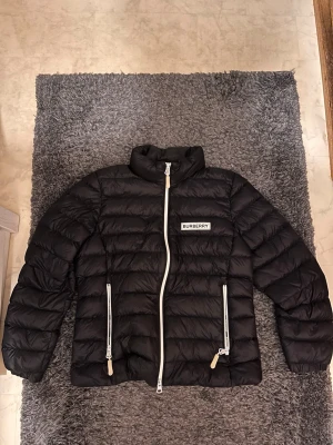 Svart dunjacka från Burberry - Snygg svart dunjacka från Burberry med vit dragkedja och två stora fickor framtill. Jackan har hög krage och Burberry-logga på bröstet. Puffig design som håller dig varm under kalla dagar. Perfekt för dig som vill ha både stil och funktion.  Dragkedjan är lite trög högst uppe vid kragen men annars är det inga fel på jackan. 
