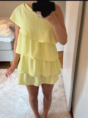 Trendig Gul one shoulder volangklänning - Säljer en ljusgul kortklänning med one shoulder och flera volanglager. Klänningen har en lös passform och är perfekt för sommardagar. Materialet känns lätt och luftigt, vilket gör den bekväm att bära. Ingen synlig logga eller märke.