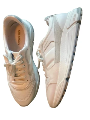 Vita sneakers från Axel Arigato - Stilrena vita sneakers från Axel Arigato med chunky sula och diskreta detaljer i mocka och mesh. Skorna är nästintill helt nya och har klassisk snörning och en modern siluett som passar till många olika outfits. Märkeslogga syns på sidan och innersulan.