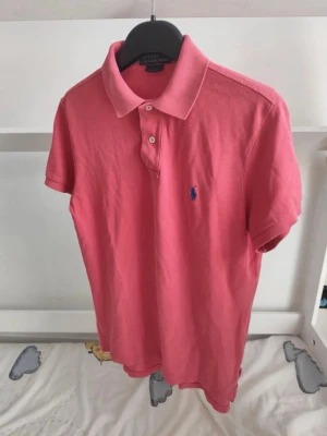 Rosa pikétröja från Ralph Lauren - Klassisk rosa pikétröja från Ralph Lauren med blå logga broderad på bröstet. Tröjan har korta ärmar, krage och knappar framtill. Tillverkad i mjuk bomull för en bekväm känsla och snygg passform.