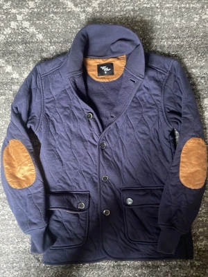Mörkblå quiltad cardigan med mockadetaljer - Snygg mörkblå cardiganjacka från Gentlemen's Concept med quiltat mönster, bruna mockadetaljer på armbågarna och två stora fickor med knappstängning. Jackan har ribbad krage och stängs med knappar framtill. Perfekt för dig som gillar klassisk men modern stil. storlek s, mycket bra skick