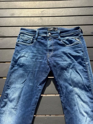 Replay anbass jeans - Säljer ett par snygga blå jeans från Replay med klassisk femficksmodell och ljusa slitningar framtill. Jeansen har normal midja, raka ben och tydliga kontrastsömmar. Perfekta för dig som gillar en avslappnad men trendig stil.