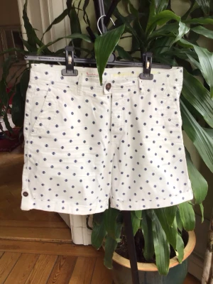 Vita prickiga shorts från Bondelid - Säljer ett par vita shorts från Bondelid med mörkblå prickigt mönster. Shortsen har normal passform, knappar i midjan och bakfickor med knappstängning. Perfekta för sommardagar och har en klassisk, fräsch look.
