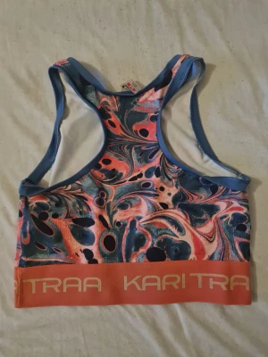 Mönstrad sport-bh från Kari Traa - Färgglad sport-bh från Kari Traa med swirl-mönster i blått, rosa och svart. Bh:n har bred resår med logga i orange och beige, samt brottarrygg och blå kantband. Perfekt för träning och aktivitet.