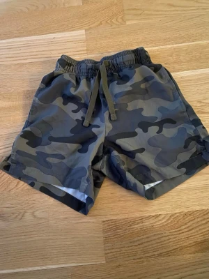 Camo shorts med snörning från Lager 157 - Snygga camouflagemönstrade shorts från Lager 157  med elastisk midja och snörning framtill. Shortsen har en loose passform och är perfekta för sommardagar. Färgerna går i grönt, svart och grått.