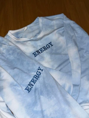 Blåvita mjukisett med tryck ENERGY för 12-13 åringar - Säljer ett par blåvita tie-dye mjukisbyxor med texten 'ENERGY' tryckt på ena benet. Byxorna har resår i midjan och vid bensluten för en skön passform. Perfekta för chill dagar hemma eller när du vill ha en avslappnad stil.