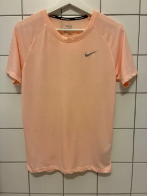 Persikofärgad Nike tränings-tshirt - Snygg  persikofärgad  tränings-tshirt från Nike med korta ärmar och rund hals. Framsidan har en diskret Nike-logga i silver och baksidan är ventilerande med små hål för extra andningsförmåga. T-shirten har dri-fit funktion. Perfekt för löpning eller gympass.