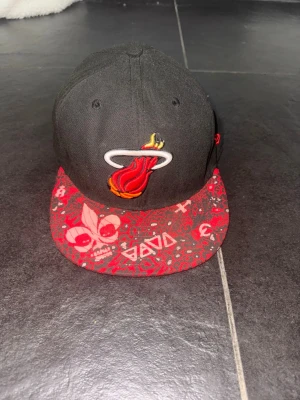 Miami Heat keps från New Era - Säljer en grå snapback-keps från New Era med Miami Heat-logga framtill. Skärmen är röd med grafiskt mönster och vita detaljer, samt coola tryck med dödskalle och symboler. Kepsen har broderad New Era-logga på sidan och är tillverkad i bomull.
