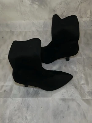 Svarta spetsiga stövletter med klack - Säljer ett par svarta stövletter med spetsig tå och smal klack. Skorna har dragkedja på sidan och är tillverkade i ett mockaliknande material. Perfekta för dig som vill ha en stilren och trendig look.