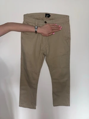 Beiga chinos från 157 - Snygga beiga chinos från 157 med klassisk rak passform. Byxorna har framfickor, bälteshällor och två bakfickor med knappdetalj. Perfekta för en clean och stilren look. Tillverkade i mjukt bomullstyg som är skönt att bära hela dagen.