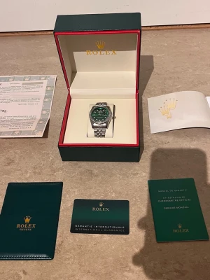 Role klocka med grön urtavla - Snygg Role klocka med grön urtavla och silverfärgat armband i metall. Klockan har en bezel med gnistrande stenar och klassisk länkdesign. Kommer i originalbox med certifikat och garantikort. Perfekt accessoar för dig som vill sticka ut.