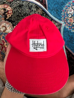 Röd keps från H&M Hybrid - Snygg röd keps från H&M med Hybrid-logga framtill. Kepsen har böjd skärm och klassisk sexpanelsdesign. Tillverkad i bomull och passar perfekt för en cool och avslappnad stil.