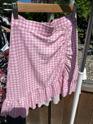 Rutig rosa kjol från Gina Tricot - Supersöt rosa och vit rutig kjol från Gina Tricot med volangkant och rynkdetalj framtill. Kjolen har en feminin och somrig look, perfekt för dig som gillar trendiga plagg med lite extra detaljer. Tillverkad i mjukt och lätt material.