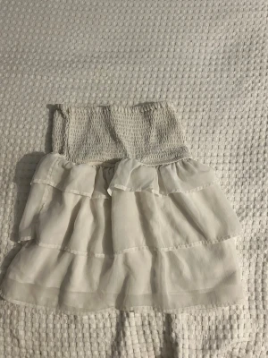Vit volangkjol från BikBok - Supersöt vit kortkjol från BikBok med bred smock i midjan och flera lager volanger. Kjolen är luftig och har en romantisk vibe, perfekt till sommaren. Tillverkad i ett lätt och mjukt material som känns skönt mot huden.
