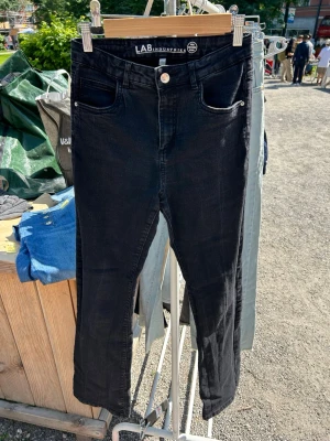 Svarta bootcut jeans från LAB Industries - Säljer ett par svarta bootcut jeans från LAB Industries med klassisk femficksdesign och knappgylf. Jeansen är tillverkade i mer hållbar denim och har en stilren look som passar till det mesta. Perfekta för dig som gillar en smal passform och enkel stil.