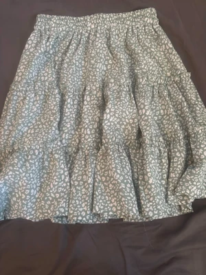 Grön volangkjol med leopardmönster - Snygg grön kjol med vita leopardfläckar och flera volanger. Kjolen har resår i midjan och ett luftigt, flowy fall som ger en avslappnad vibe. Perfekt för dig som gillar djurmönster och vill ha något bekvämt men ändå trendigt.
