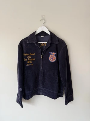 Mörkblå/Lila FFA Jacka - Äkta vintage FFA-jacka (Future Farmers of America) från Illinois (97/98). Den har tillhört Stephen Leavell, vilket gör den till ett unikt samlarobjekt med en personlig historia bakom sig!  Vintagepatina med slitage lite här och var, främst på ärmen (se sista bilden) vilket jag dragit av för på priset. Passformen är boxig och cropped, ca S i storlek.