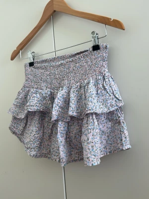 Blommig volangkjol med smock - Supersöt kort kjol med blommigt mönster i pastellfärger. Kjolen har bred smock i midjan och dubbla volanger som ger en luftig och lekfull look. Perfekt för varma dagar och enkel att matcha med en enkel topp.
