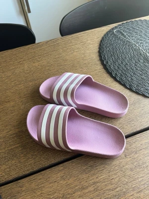 Ljuslila Adidas badsandaler - Säljer ett par ljuslila Adidas badsandaler med klassiska vita ränder över foten. 