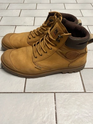 Palladium Kängor - Snygga ljusbruna kängor från Palladium med robust sula och högt skaft. Ovandelen är i läder och textil, med vadderad brun krage för extra komfort. Klassisk logotyp på sidan och tåliga snörhål i metall. Perfekta för höst och vinter.