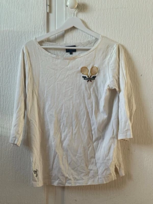 Vit Båstad Tennis t-shirt från SIR - Vit t-shirt från SIR med tryck av två tennisracketar och texten 'Båstad Tennis' på ryggen. Trekvartsärm och rund halsringning. Tillverkad i mjuk bomull, perfekt för en sportig och avslappnad look.