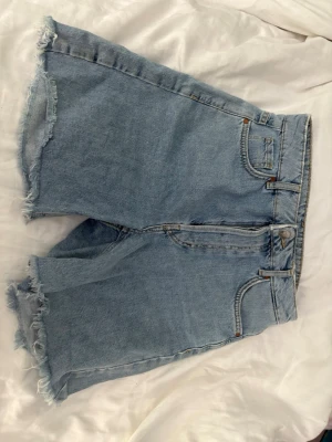 Ljusblå denimshorts med rå kant - Säljer ett par ljusblå denimshorts från bikbok med hög midja och råa, fransiga benslut. Når precis över knäna. Klassisk femficksmodell med dragkedja och knapp framtill. Perfekta för sommaren och ger en avslappnad vibe.
