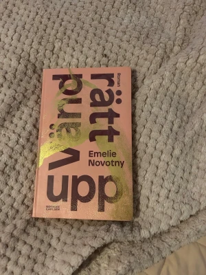 Vänd rätt upp av Emelie Novotny - Romanen 'Vänd rätt upp' av Emelie Novotny har ett rosa hårdband med stora lila bokstäver och gulddetaljer på omslaget. Boken är tryckt på papper och har en modern, ungdomlig design. Perfekt för dig som gillar samtida svensk skönlitteratur.