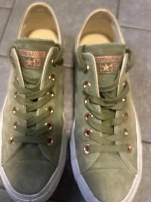 Gröna Converse All Star sneakers - Snygga gröna Converse All Star sneakers i mocka med vita sulor och guldfärgade metalldetaljer. Klassisk låg modell med matchande gröna skosnören och logga på plösen. Perfekta för dig som vill ha en stilren och trendig look.