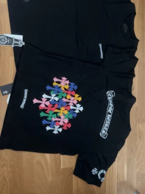Svart t-shirt med färgglada blommor - Säljer en svart t-shirt från Chrome Hearts med ett stort tryck av färgglada blommor på framsidan. T-shirten har korta ärmar och vita logodetaljer på bröstet och ärmen. Perfekt för dig som gillar streetwear och unika prints.